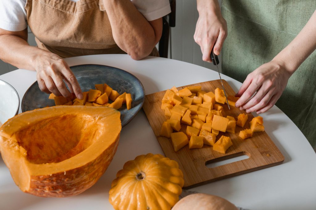 Hands cutting diced squash on a wooden board with a halved squash and plated pieces nearby.
